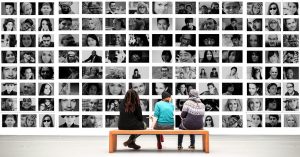 Three people sitting on a bench looking at a wall filled with portraits of a diversity of people.